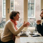 Vrouw gebaart expressief tijdens gesprek in Amsterdams café met bakstenen muren en grote ramen, koffiekopjes op houten tafel
