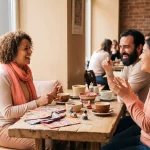 Drie mensen in geanimeerd gesprek aan houten cafétafel met kleurrijke stoffen en culturele voorwerpen, Amsterdam