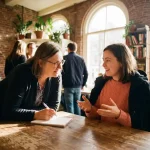 Onderzoeker met notitieboek in gesprek met jonge vrouw aan houten café tafel in Amsterdam, warme middag licht