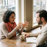 Twee mensen in gesprek aan een houten cafétafel in Amsterdam, één persoon gebaart expressief, ander luistert aandachtig, koffiekopje en notitieboek erbij.