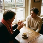 Twee mensen in gesprek aan een houten cafétafel in Amsterdam, met zacht natuurlijk licht en een koraalrode sjaal als accent.