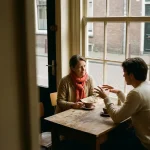 Twee mensen in gesprek aan een versleten houten cafétafel in Amsterdam, met keramische koffiekopjes en zacht natuurlijk daglicht.