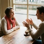 Twee mensen in gesprek aan een houten cafétafel in Amsterdam, één gebaart terwijl de ander aandachtig luistert, zachte daglichtsfeer.
