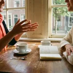 Twee mensen in levendig gesprek aan een versleten houten cafétafel in Amsterdam, met koffiekopjes en een notitieboekje erbij.