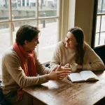 Twee mensen in gesprek aan een houten cafétafel in Amsterdam, één gebarend, de ander aandachtig luisterend, met koffie en notitieboek.