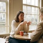 Vrouw in gesprek aan een houten cafétafel in Amsterdam, handen gebarend, zachte daglichtsfeer met koraalrood keramisch kopje.
