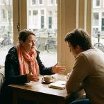 Klant en consultant in gesprek aan een houten cafétafel in Amsterdam, met open handgebaren en aandachtige houding bij zacht daglicht.