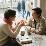 Twee mensen in gesprek aan een versleten houten cafétafel in Amsterdam, met koffiekopjes en een open notitieboek.