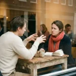 Twee mensen in gesprek aan een houten cafétafel in Amsterdam, met koffie en een open notitieboek, zachte daglichtsfeer.