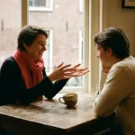 Twee mensen in gesprek aan een versleten houten cafétafel in Amsterdam, warm natuurlijk licht, keramische koffiekop op de voorgrond.
