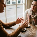 Twee mensen in gesprek aan een verweerde houten cafétafel in Amsterdam, één met expressieve handgebaren, de ander luisterend met een fronsende blik.