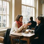 Vrouw in levendig gesprek aan een houten cafétafel in Amsterdam, met keramische koffiekopjes en zacht natuurlijk raamlicht.