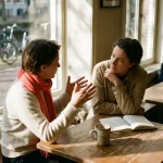 Twee mensen in gesprek aan een houten cafétafel in Amsterdam, met een koffiekopje en open notitieboek in warm natuurlijk licht.