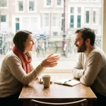 Twee mensen in gesprek aan een versleten houten cafétafel in Amsterdam, één leunt voorover met open handen terwijl de ander aandachtig luistert.