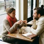 Twee mensen in gesprek aan een versleten houten cafétafel in Amsterdam, met koffie en een open notitieboek.