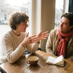 Twee mensen in gesprek aan een versleten houten cafétafel in Amsterdam, met een keramische koffiekop en notitieboekje erbij.