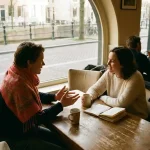 Twee mensen in gesprek aan een houten cafétafel in Amsterdam, met een koffiekopje en notitieboekje, in warm natuurlijk daglicht.