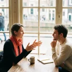 Twee mensen in gesprek aan een versleten houten cafétafel in Amsterdam, één leunt voorover met open handen terwijl de ander aandachtig luistert.