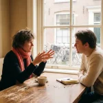 Twee mensen in gesprek aan een versleten houten cafétafel in Amsterdam, met koffiekopje en notitieboekje, warme daglichtsfeer.