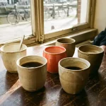 Handgemaakte keramische koffiekopjes op een versleten houten cafétafel in Amsterdam, met een koraalrood kopje als blikvanger in zachte middagzon.